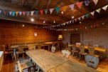 Innenraum des Veranstaltungsortes "Hüfnerhaus". Mehrere Tische und Stühle aus Holz stehen im Raum. Von der Decke hängen dekorativ mehrere Wimpelketten.